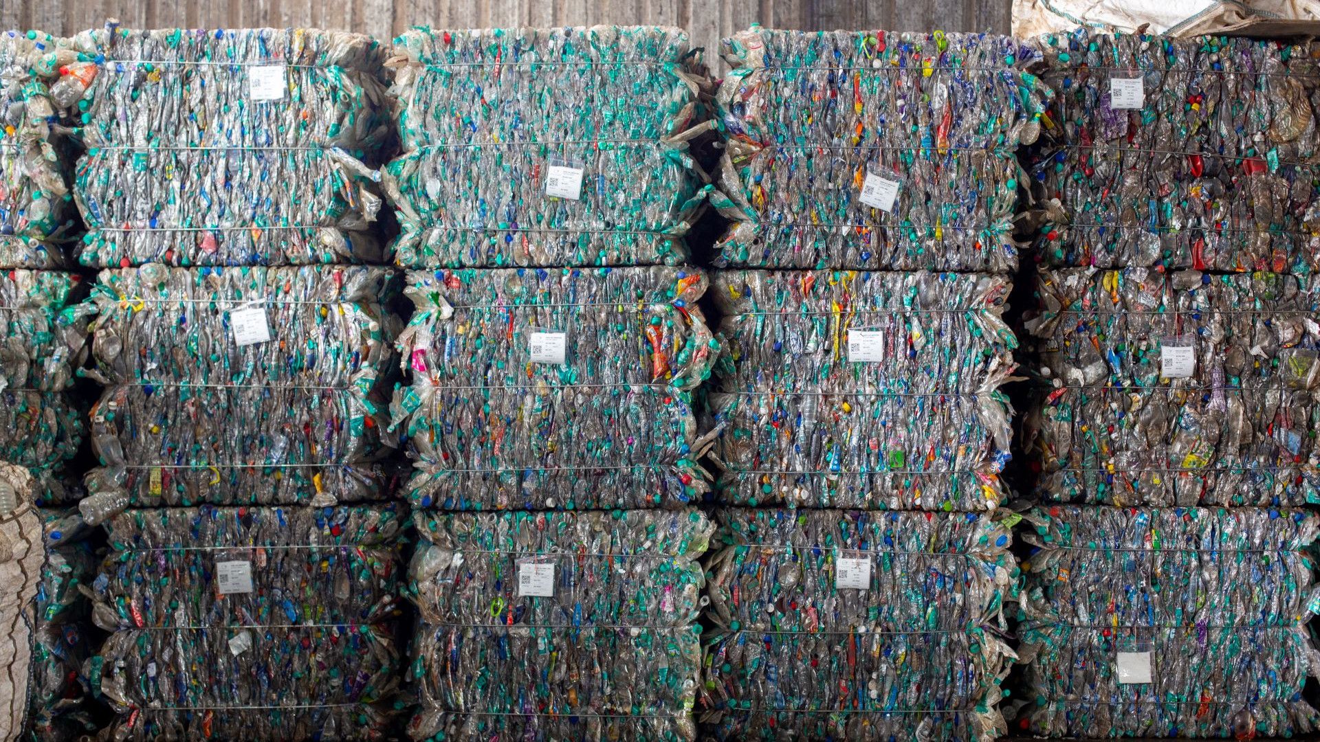 Stacks of compressed bales of plastic waste, bound tightly and arranged in rows. The bales consist of various colourful plastic materials.