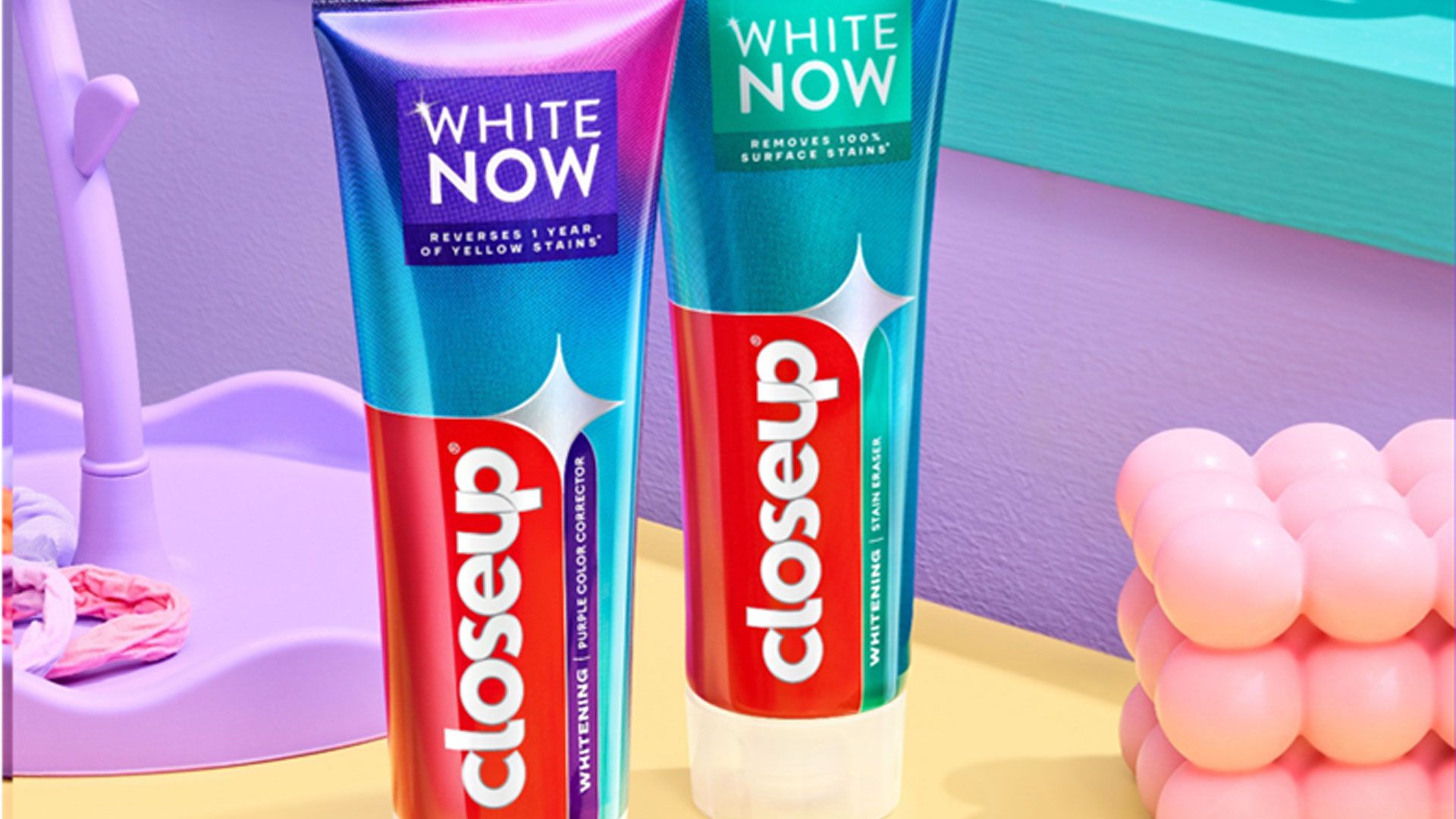 Two tubes of Closeup White Now toothpaste standing on a pastel bathroom counter with a pink toothbrush in the foreground.