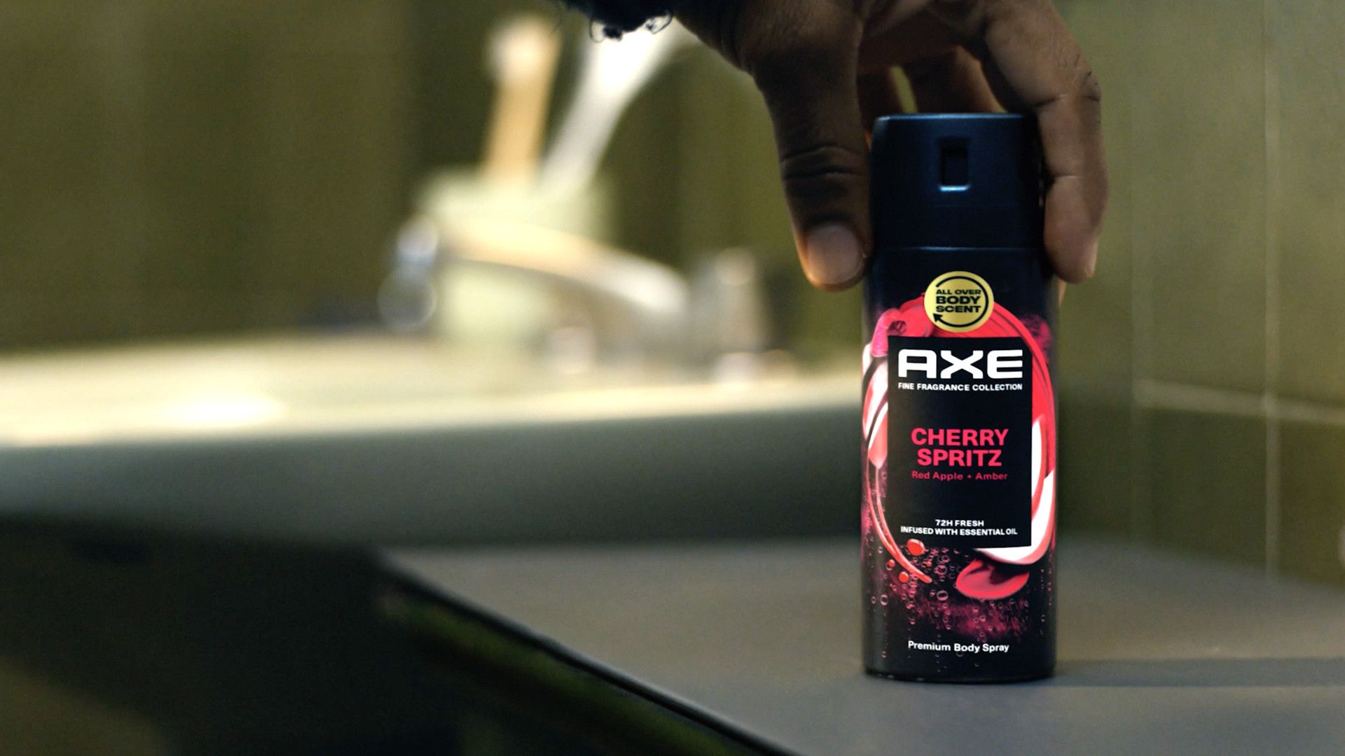 A close-up of a person’s hand placing a black can of AXE Cherry Spritz body spray on a bathroom counter. The can features a red and white swirl design. The blurred sink and tiles in the background create a casual bathroom setting.