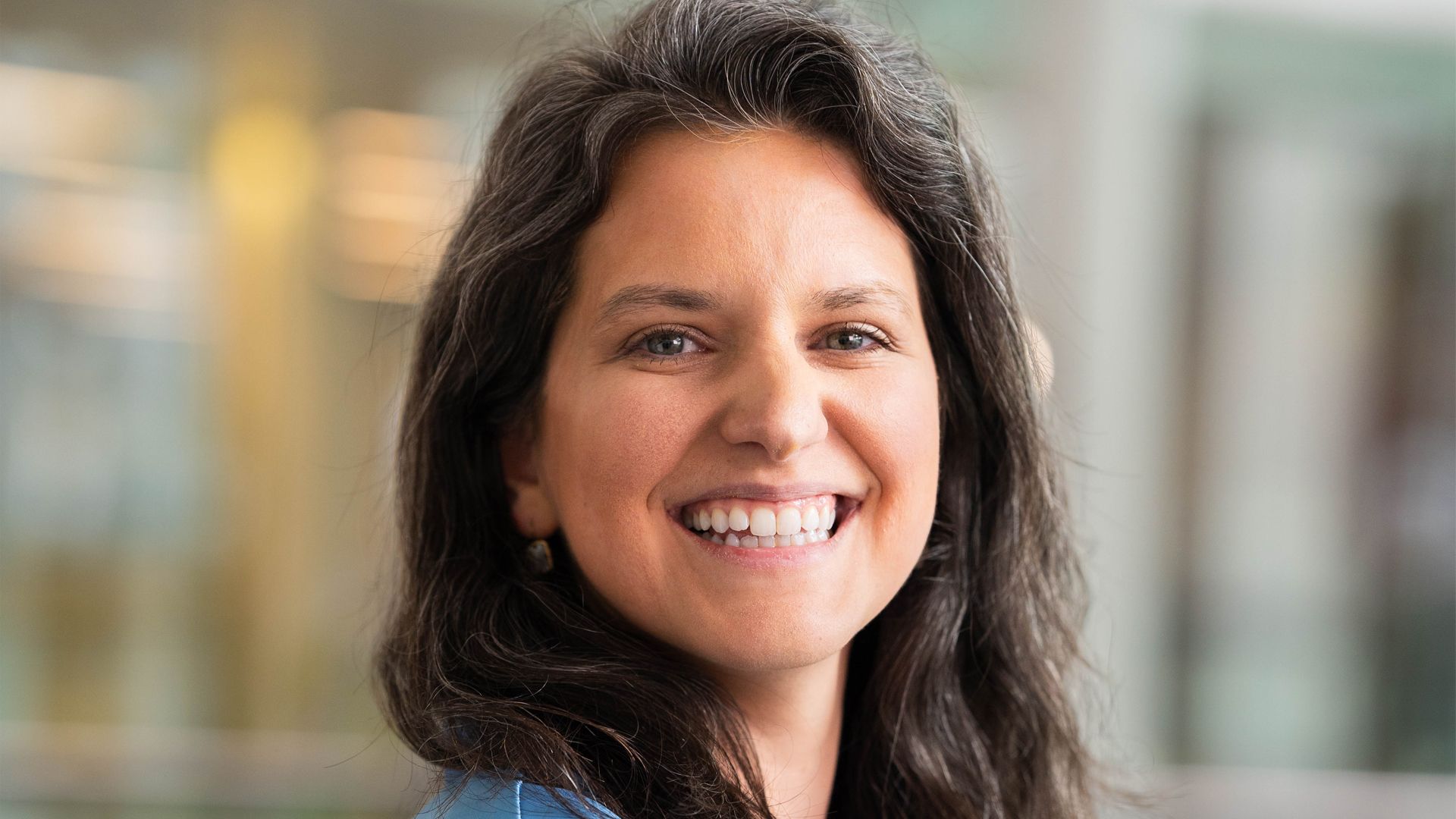 A person with long, dark hair and a broad smile in an indoor setting.