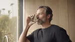 Person indoors holding a glass near their mouth, wearing a black shirt, with a blurred view of greenery outside the window.