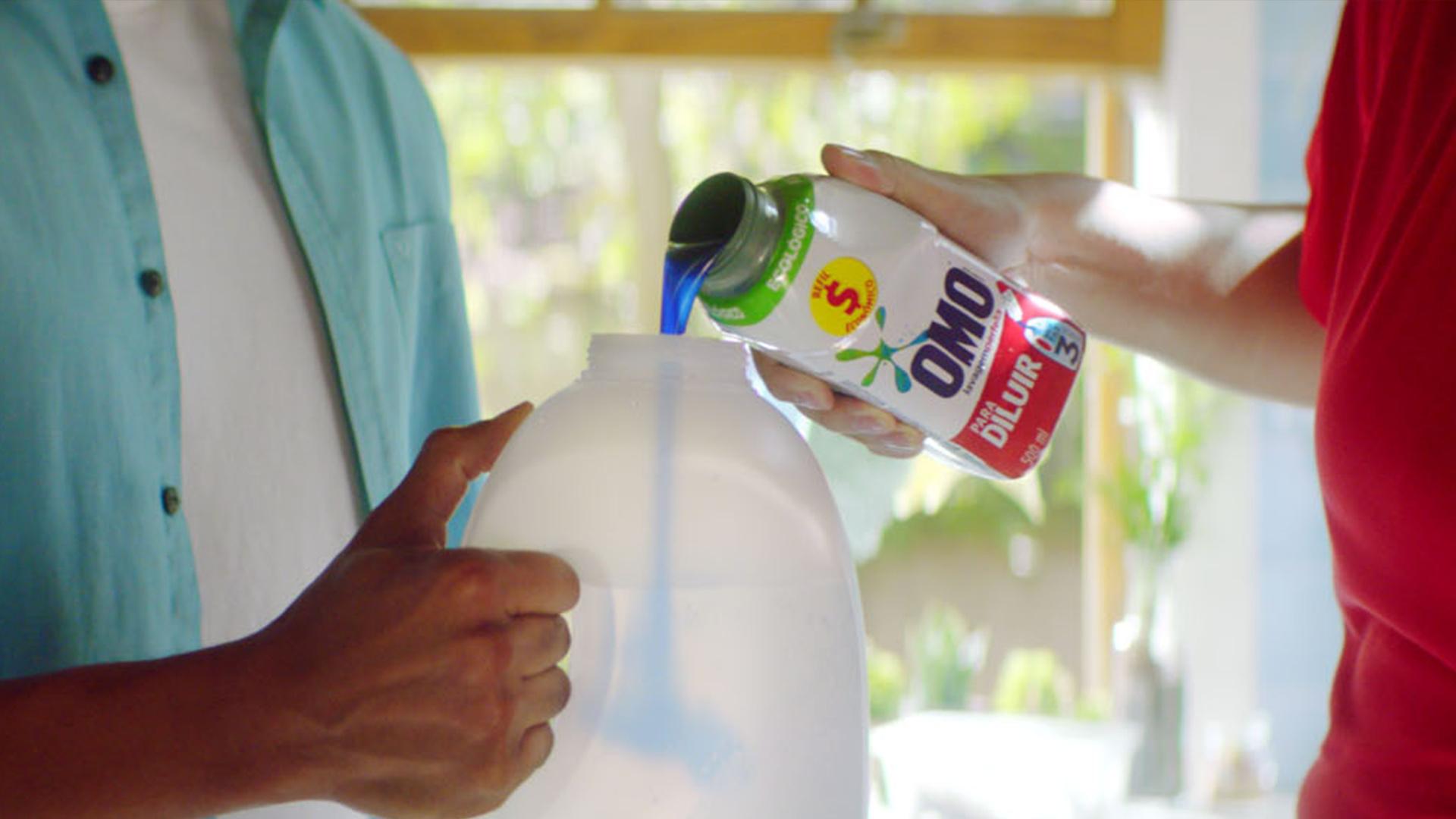 A close-up of a person pouring blue liquid detergent from an OMO-branded bottle into a larger white plastic container which a second person is holding.