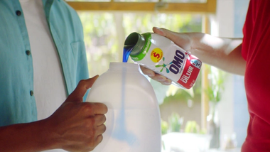 A close-up of a person pouring blue liquid detergent from an OMO-branded bottle into a larger white plastic container which a second person is holding.