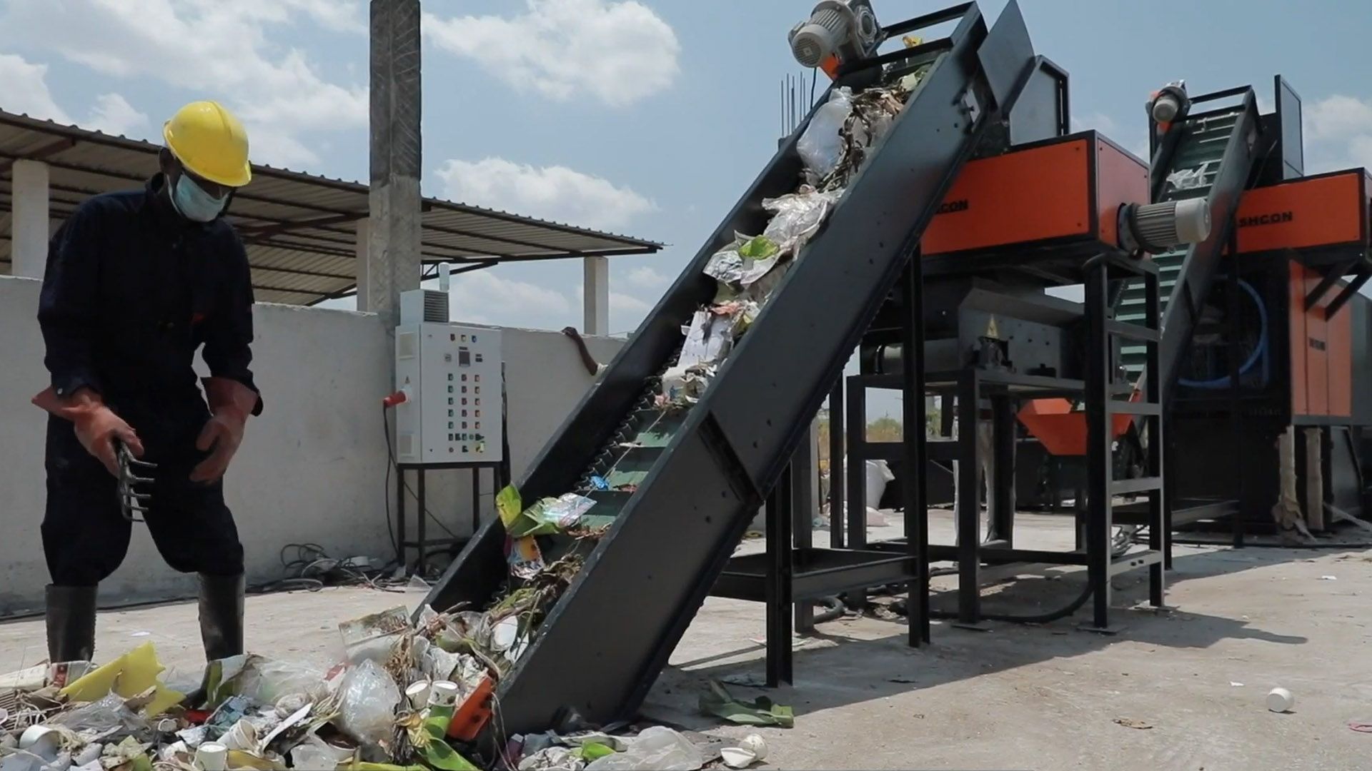 Male worker in overalls and hard hat in a yard, loading waste onto the conveyor belt of a TrashCon sorting and recycling machine.