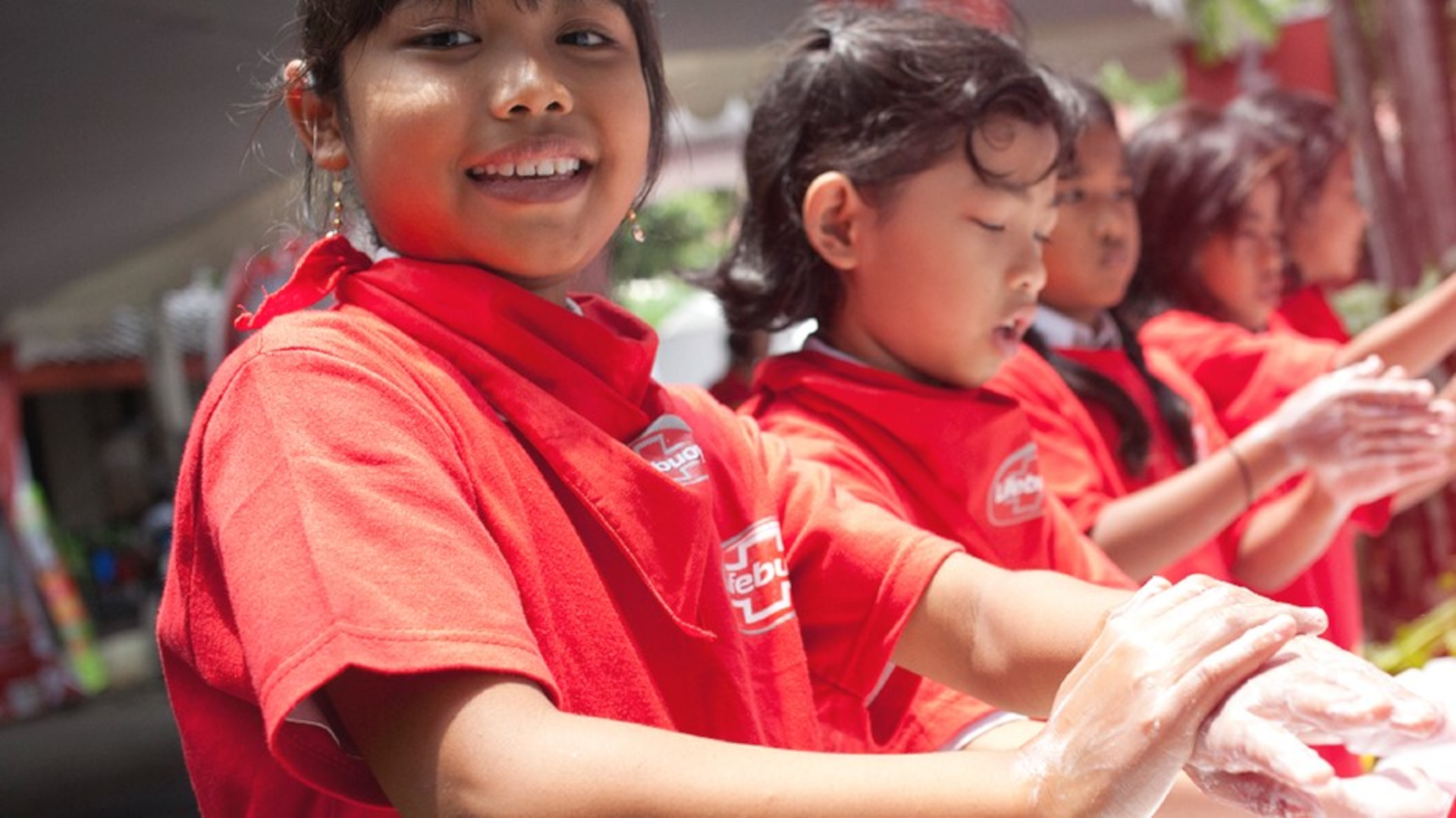 Smiling children wash their hands with Lifebuoy soap. They are wearing red T-shirts printed with a Lifebuoy logo.