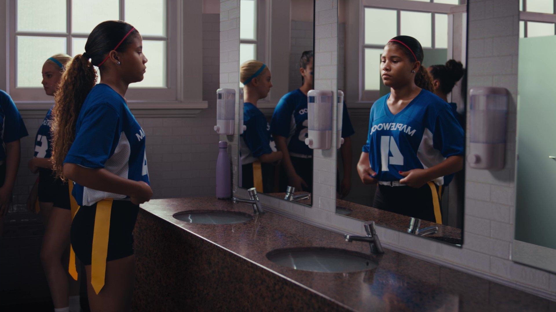 A group of girls in sports uniforms stand in a locker-room–style bathroom with sinks and mirrors. One girl adjusts her uniform while others stand nearby, reflected in the mirrors.