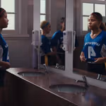 A group of girls in sports uniforms stand in a locker-room–style bathroom with sinks and mirrors. One girl adjusts her uniform while others stand nearby, reflected in the mirrors.