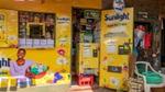 A small storefront featuring bright yellow branding for ‘Sunlight’ laundry detergent. The store includes a detergent vending machine and a small service counter, and is adorned with colourful illustrations of flowers.