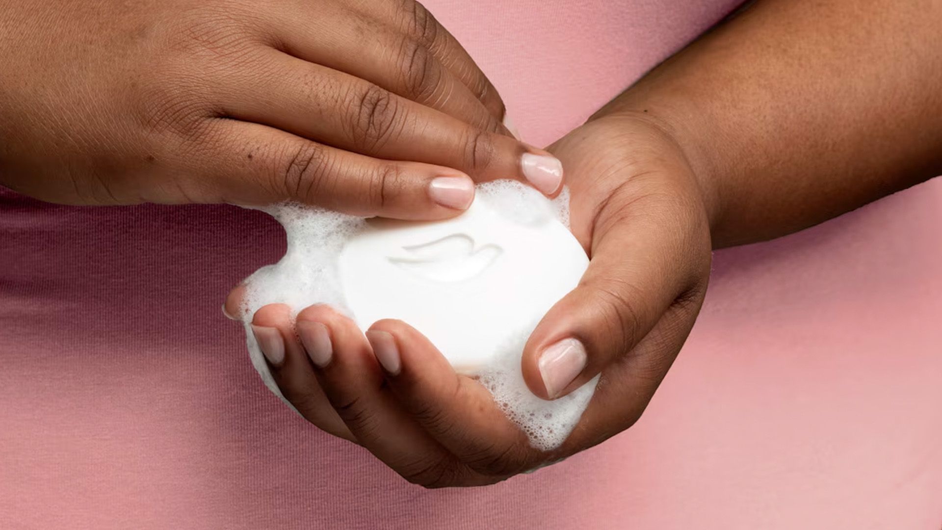 A closely cropped photo of a person in a pink top holding a Dove soap bar, with frothy white bubbles on their hands.