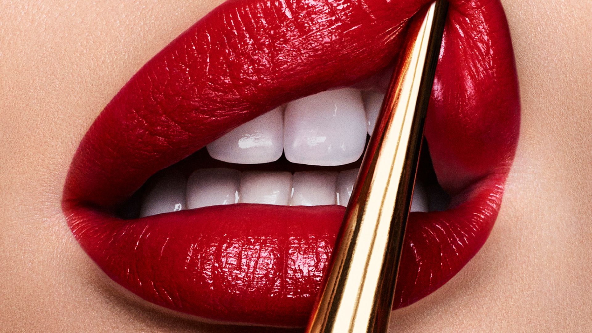 Extreme close-up of glossy red lips with white teeth, applying lipstick using a gold applicator.
