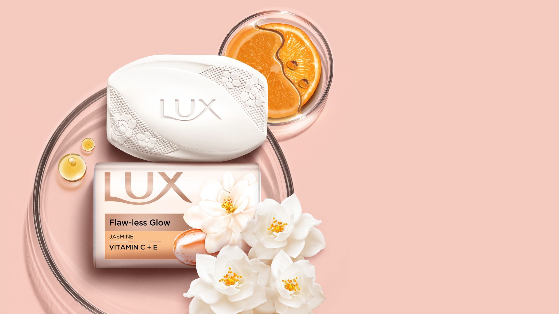 Two bars of LUX soap – one packaged, one not – from the Flaw-less Glow range are on the left of the image. Several jasmine flowers sit alongside the soap. This all sits on a pink beige background.