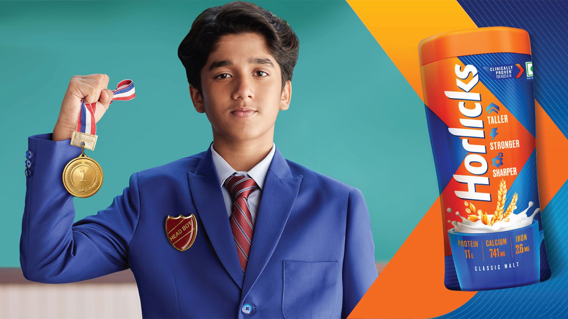 A schoolboy wearing a blue blazer with a ‘Head boy’ badge pinned to it stands with his right arm bent, fist clenched, grasping a 1st place medal. To the right of him is a jar of Horlicks.