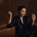 Woman sat on a brown couch dressed in black with a beauty product in her hand.