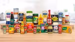 A selection of well known food and condiment brands, including Hellmann’s mayonnaise, Frank’s RedHot, Cholula Hot Sauce, Knorr seasonings, French’s mustard, spices and condiments, arranged side by side on a wooden table.