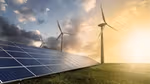 Solar panels installed on a grassy field with wind turbines in the background under a partly cloudy sky and bright sunlight.