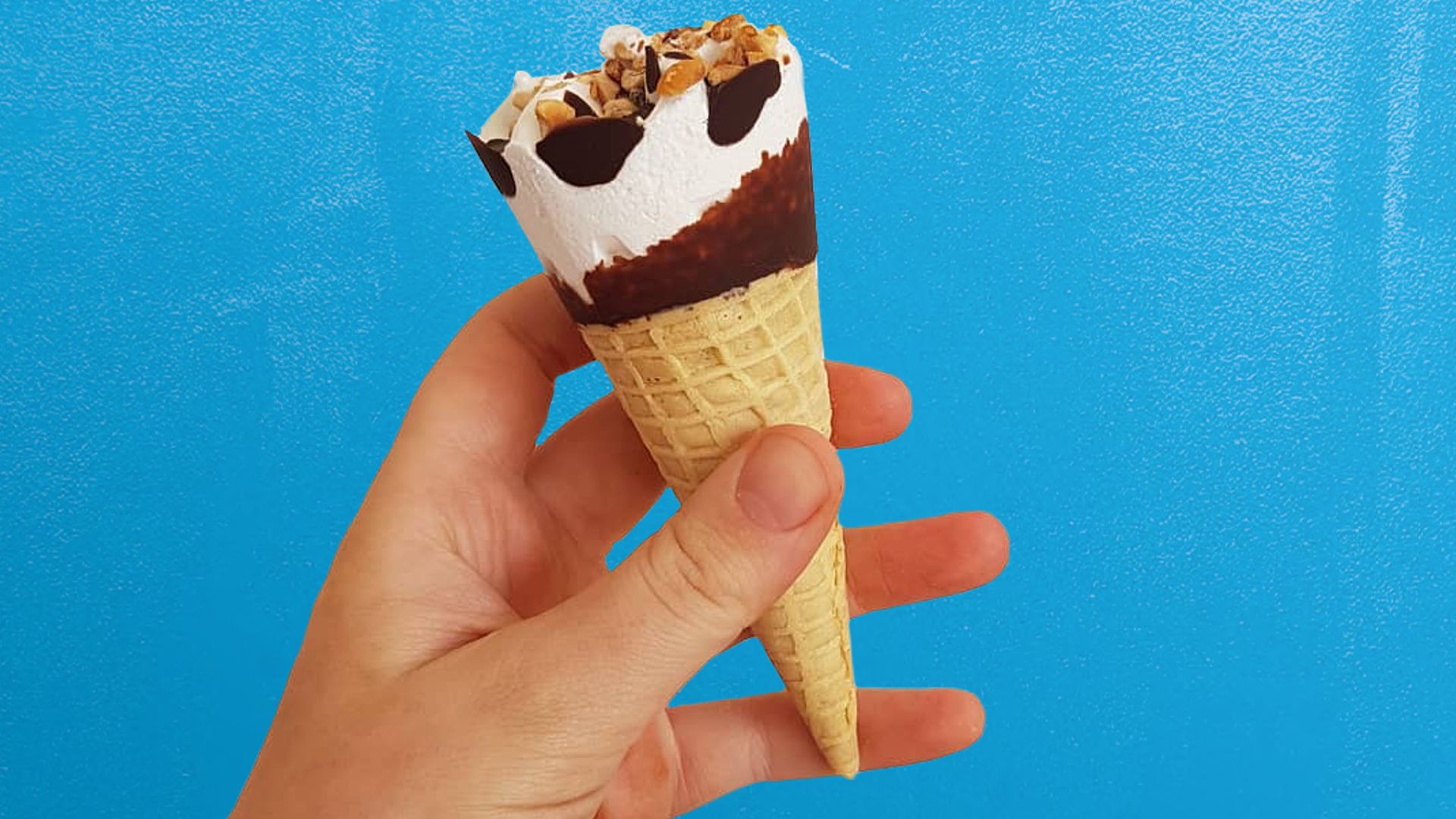 A hand holding a Cornetto ice cream in blue and white packaging. Topping is chocolate and nut. Set on a blue background.
