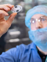 A gloved hand holding a small, circular plastic component in a cleanroom environment with machinery in the background.