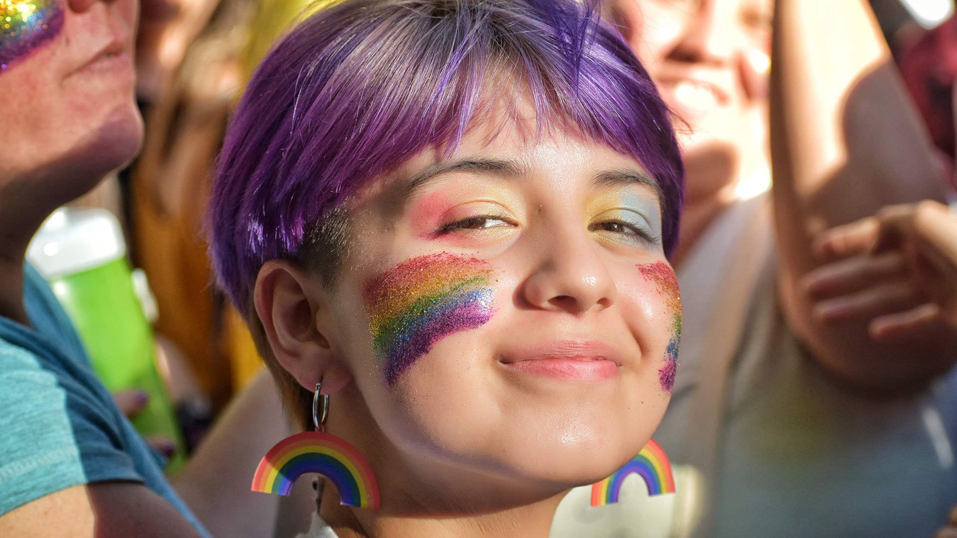 Someone smiling among a group of people with a rainbow-painted face and rainbow earrings.
