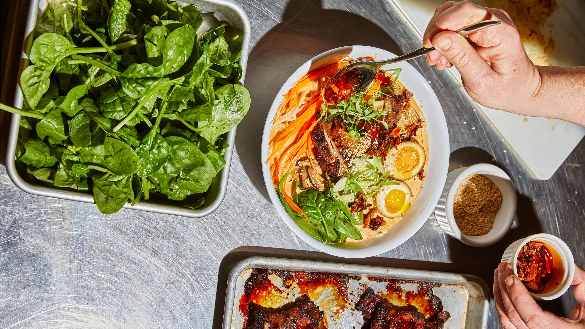 An image of dishes on a stainless-steel table, including a tray of green leaves and a ramen bowl over which red sauce is being drizzled.