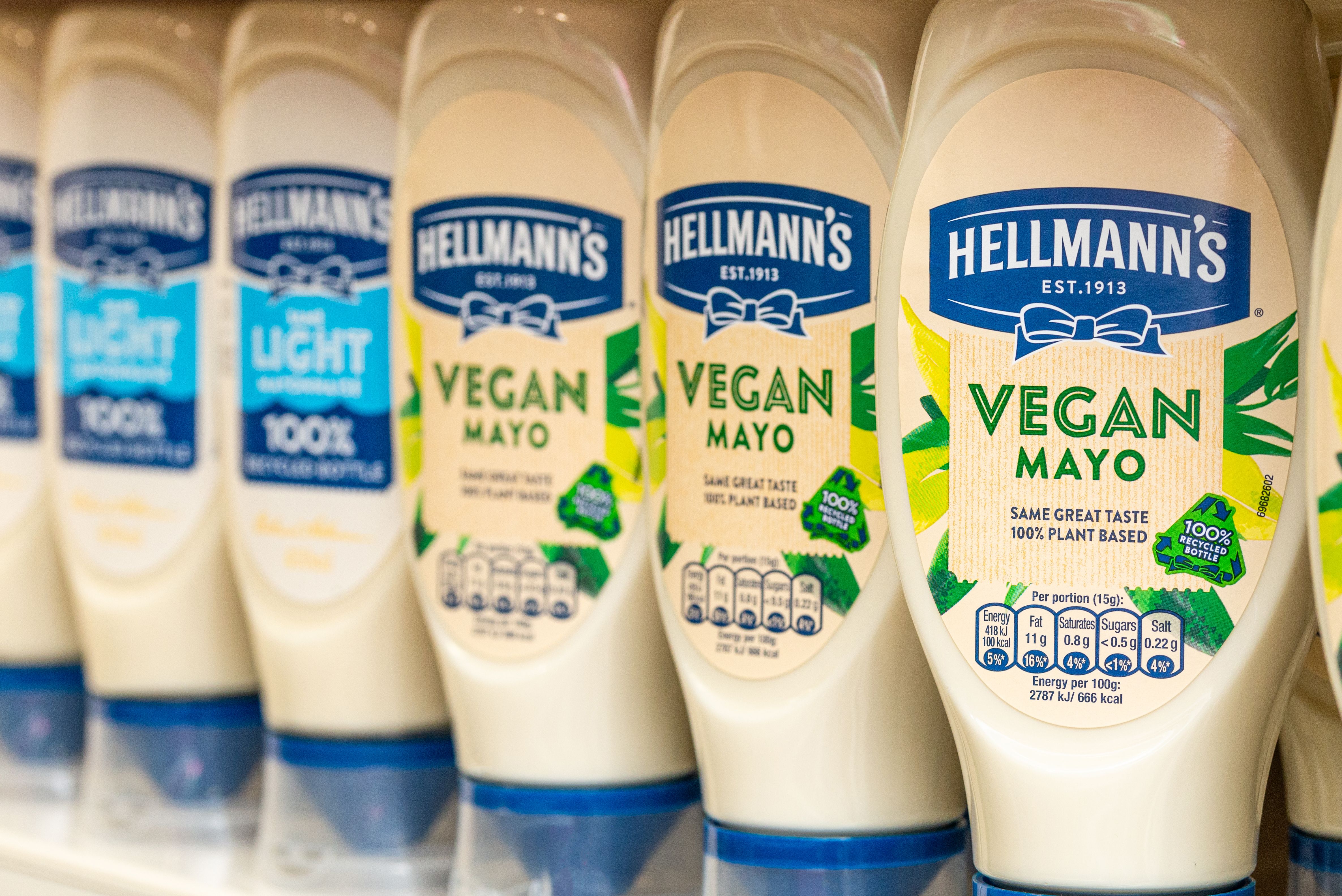 A close-up of Hellmann’s mayonnaise bottles lined up on a shelf, featuring a mix of ‘Light’ and ‘Vegan Mayo’ varieties. The Vegan Mayo bottles are labelled with ‘Same Great Taste, 100% Plant Based’ and ‘100% Recycled Bottle’. 