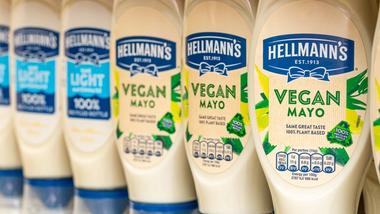 A close-up of Hellmann’s mayonnaise bottles lined up on a shelf, featuring a mix of ‘Light’ and ‘Vegan Mayo’ varieties. The Vegan Mayo bottles are labelled with ‘Same Great Taste, 100% Plant Based’ and ‘100% Recycled Bottle’.