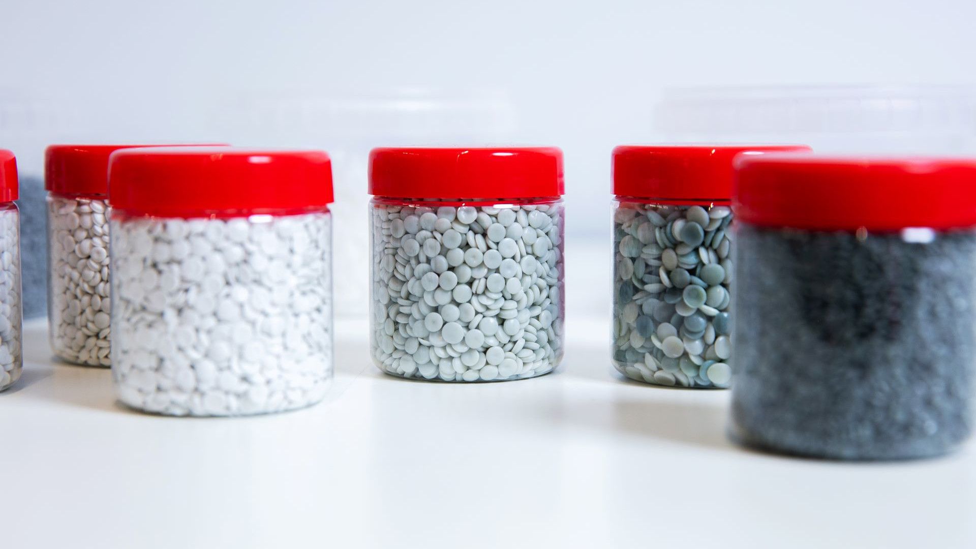 Several transparent jars with red lids containing small plastic pellets in various shades, arranged on a white surface.