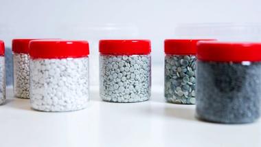 Several transparent jars with red lids containing small plastic pellets in various shades, arranged on a white surface.