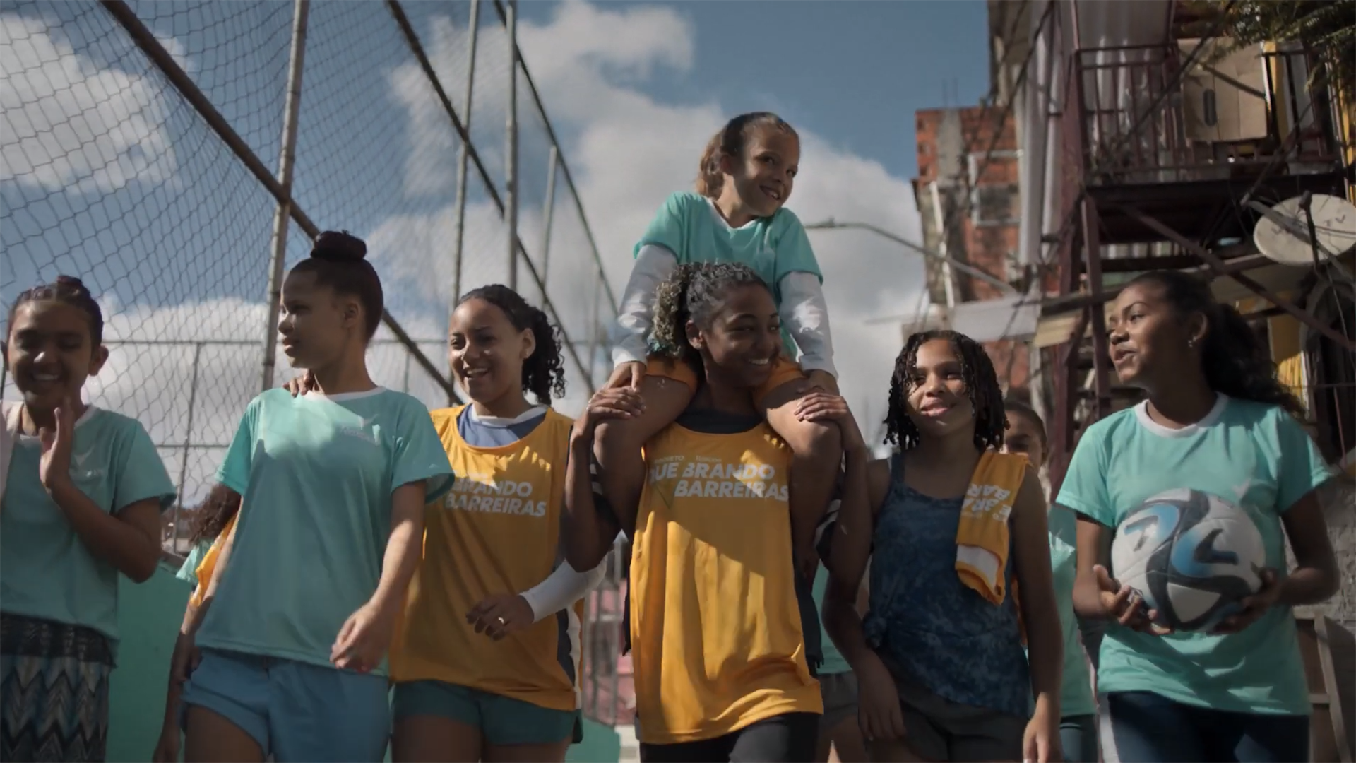 7 girls are between a fenced football pitch and buildings. Six are walking with one of them carrying a football. The smallest girl is being carried on the shoulders of the girl in the middle. 