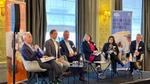 Six panellists seated on a stage at an event, with banners in the background displaying the text 'Creating a clean future – without compromise.'