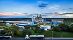 Industrial site with tanks, buildings, and a tanker truck on a road, set against a blue sky with clouds and greenery.