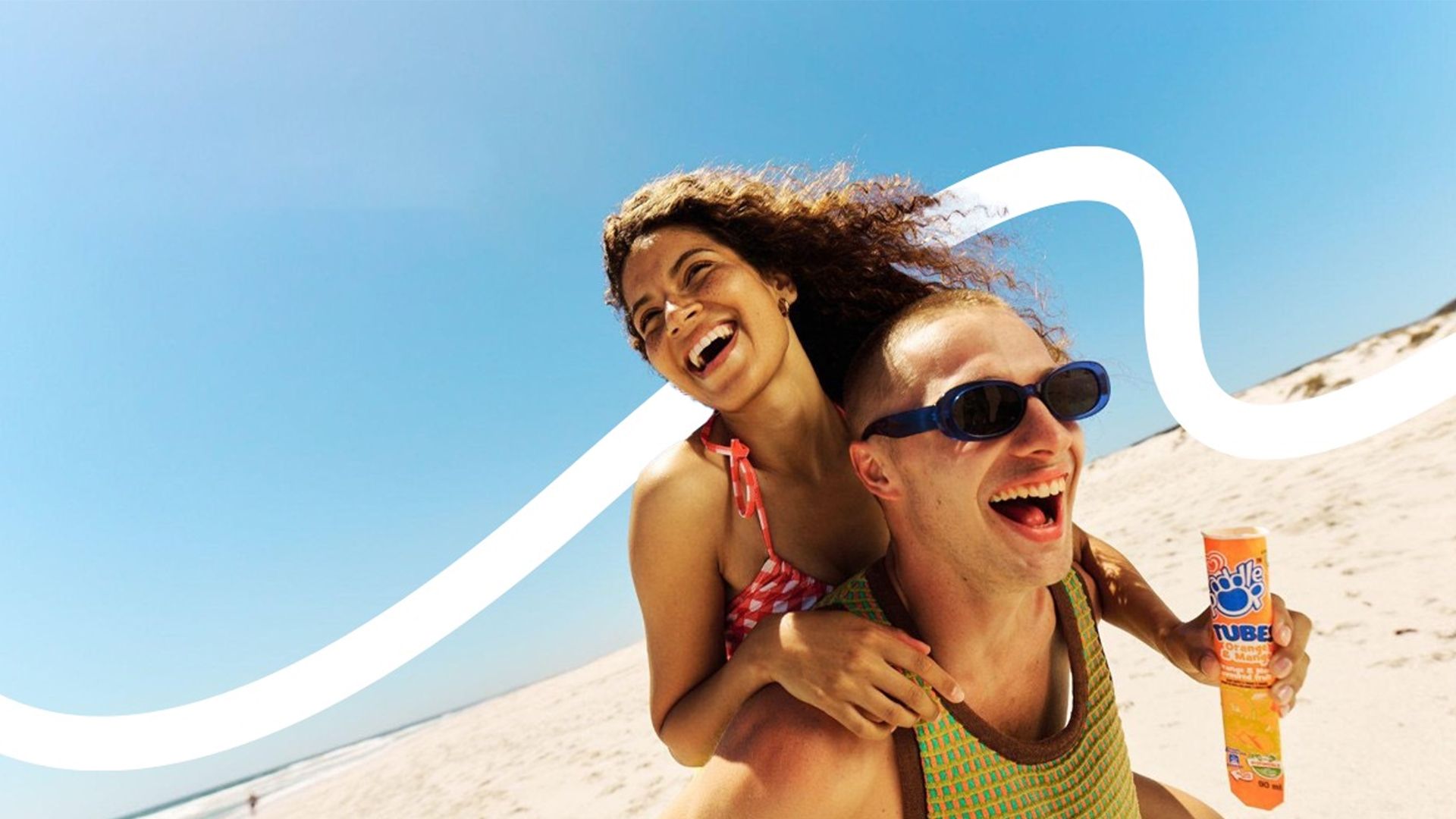 Two people enjoying a sunny day at the beach. One is giving the other a piggyback ride. The person being carried wears a red and white checked swimsuit and has curly hair, while the other has short hair, sunglasses, and holds an orange Paddle Pop. A clear blue sky with a white wavy graphic overlays the background.