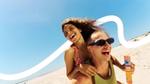 Two people enjoying a sunny day at the beach. One is giving the other a piggyback ride. The person being carried wears a red and white checked swimsuit and has curly hair, while the other has short hair, sunglasses, and holds an orange Paddle Pop. A clear blue sky with a white wavy graphic overlays the background.