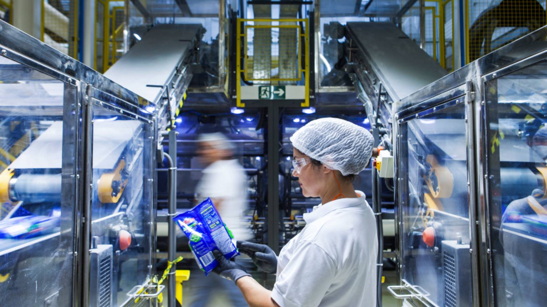 A factory worker looks at some packaging. Factory machinery and factory workers are blurred in the background.