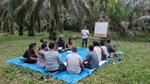 A photograph of a group of people seated in a circle on a blue tarpaulin sheet surrounded by palm trees. A facilitator is conducting a training session.