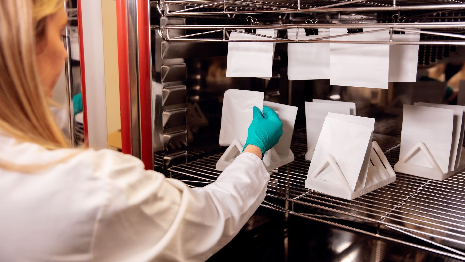 A packaging expert working in a lab testing paper-based flexible packaging samples. The packaging expert is a woman with blonde hair. She is wearing a white lab coat and turquoise gloves, and she is holding one of the samples.