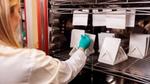 A packaging expert working in a lab testing paper-based flexible packaging samples. The packaging expert is a woman with blonde hair. She is wearing a white lab coat and turquoise gloves, and she is holding one of the samples.
