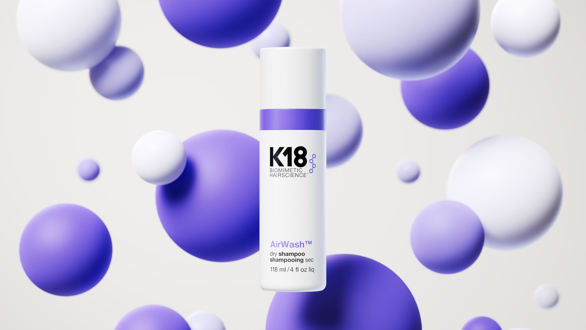 A white bottle of K18 AirWash dry shampoo stands centred against a soft background filled with floating white and purple spheres of different sizes. The clean, minimalist packaging features black and purple text, while the surrounding spherical shapes create a light, airy feeling.