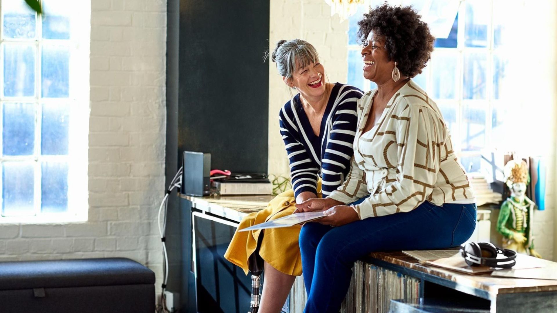 Two women sitting on a worktop chatting and laughing.