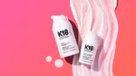 Two products from the K18 range – both white plastic bottles – lying on a bright pink background with a swirl of he product smeared on it.