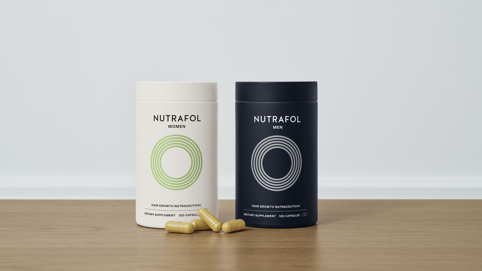 Two cylindrical Nutrafol supplement containers stand side by side on a wooden surface against a simple, pale background. The container on the left is cream-coloured and labelled for Women, while the one on the right is dark navy and labelled for Men. Each has a circular, line-art style design on the front. Several beige capsules lie scattered in front of the containers.