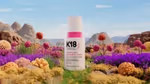 Bottle of K18 HeatBounce conditioning heat protectant displayed in a vibrant, colourful landscape with rocky mountains in the background and clusters of bright, spherical plants in shades of purple, pink, and yellow in the foreground.