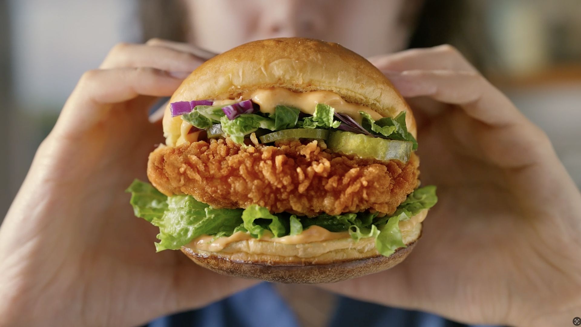 Close-up of a crispy fried chicken sandwich being held with both hands. The sandwich features a golden-brown breaded chicken fillet, fresh lettuce, pickles, red onion slices, and a creamy sauce inside a toasted bun.
