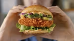 Close-up of a crispy fried chicken sandwich being held with both hands. The sandwich features a golden-brown breaded chicken fillet, fresh lettuce, pickles, red onion slices, and a creamy sauce inside a toasted bun.