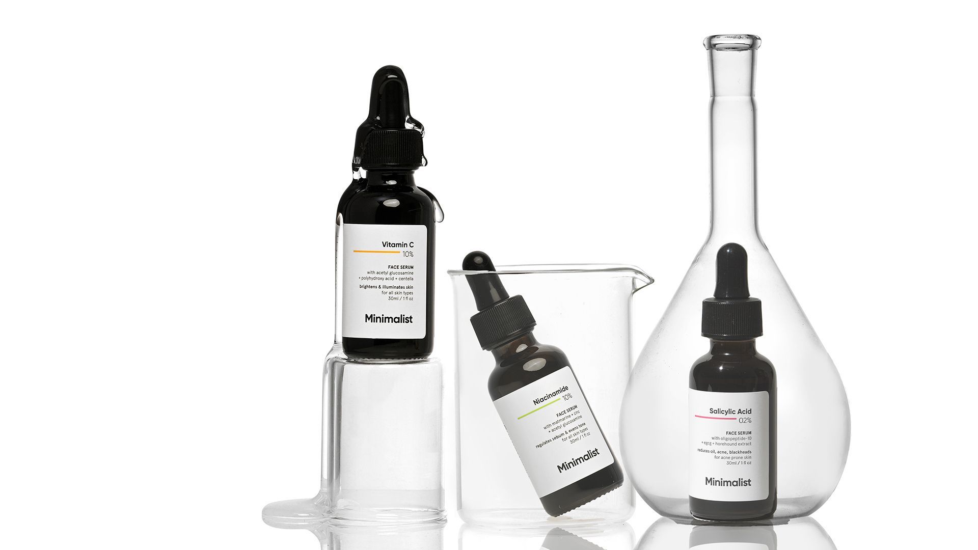 Three brown bottles of serums. One is placed on an upturned glass jugs, another is inside a glass jar and the third is inside a glass bottle.   