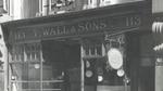 Black and white photograph of the storefront of T. Wall & Sons Ltd at number 113. The store name is displayed above the entrance, flanked by the number 113 on both sides. A hanging sign with partially legible text is visible in front of the store. Large display windows reveal various items inside.