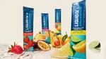 A variety of Liquid I.V. Hydration Multiplier electrolyte sachets in different flavours, including Strawberry and Lemon Lime. The packets are arranged around fresh fruits like strawberries, passion fruit, pineapple, lemon, lime, and orange slices on a light-coloured surface with water droplets.