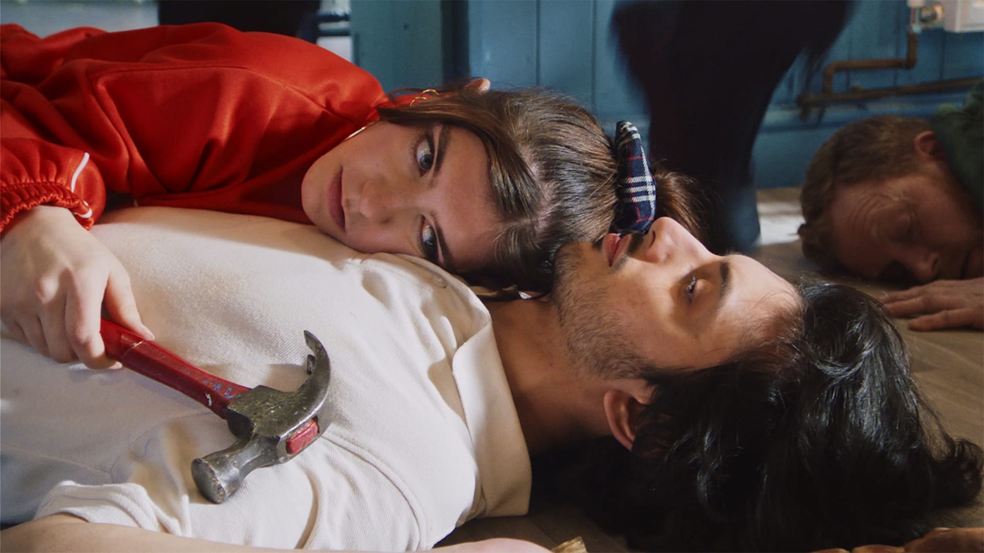A man lying on his back on the floor. A woman is also lying down and hugging him while holding a hammer in her hand.