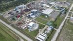 An aerial view of an industrial facility featuring buildings, large storage tanks and some green spaces, surrounded by roads and trees in the background.