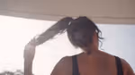 Back view of a person holding their ponytail in bright sunlight with lens flare, wearing a sleeveless top.