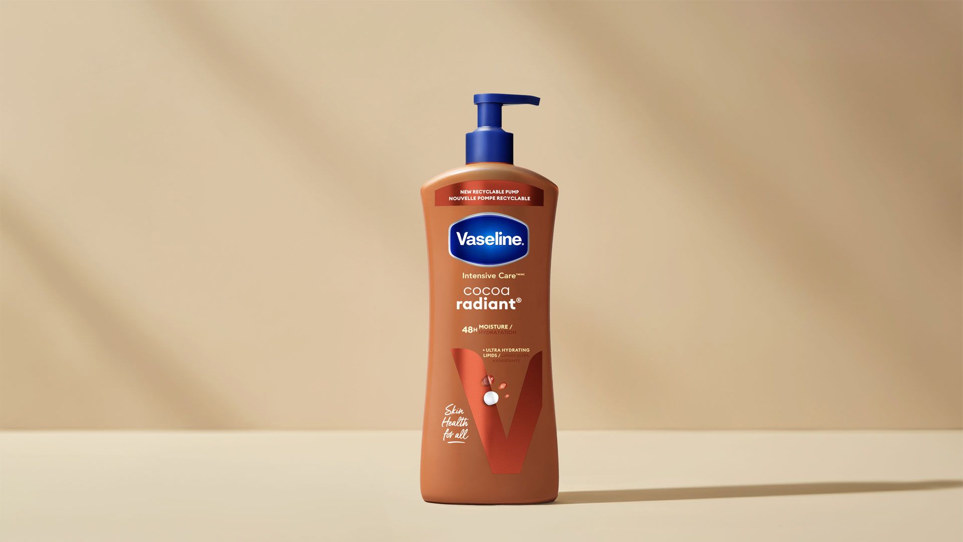 A tall, brown bottle of Vaseline Intensive Care Cocoa Radiant lotion standing upright against a softly lit beige background. The bottle has a deep blue pump at the top and a subtle gradient shadow beneath it. The front label displays the Vaseline logo and text describing the product’s moisturising properties. The scene is minimalistic, with warm lighting creating gentle shadows.